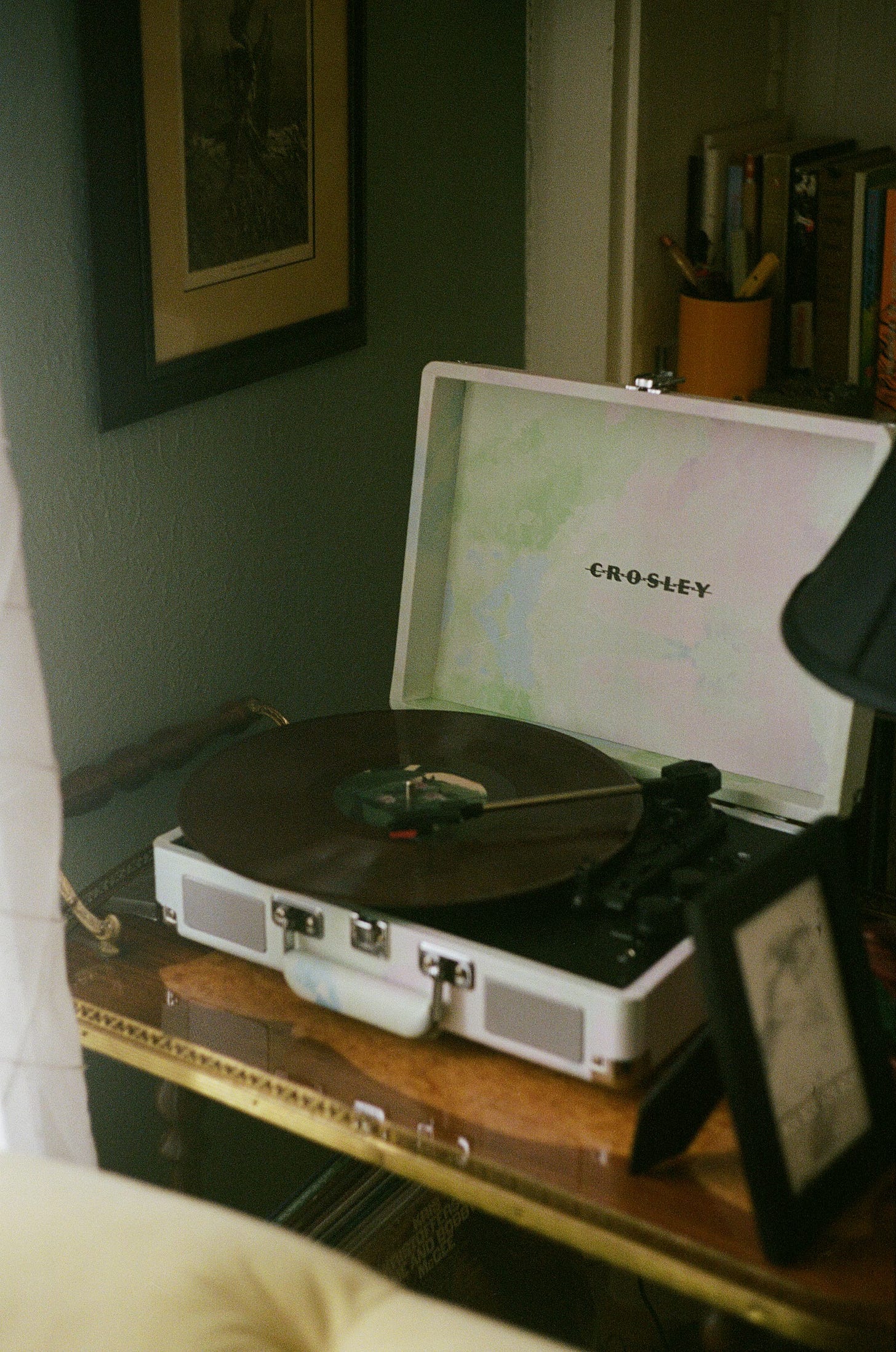 A white Crosley portable record player with its lid open, showing a vinyl record on the turntable, placed on a wooden shelf against a green wall with a framed picture in the background