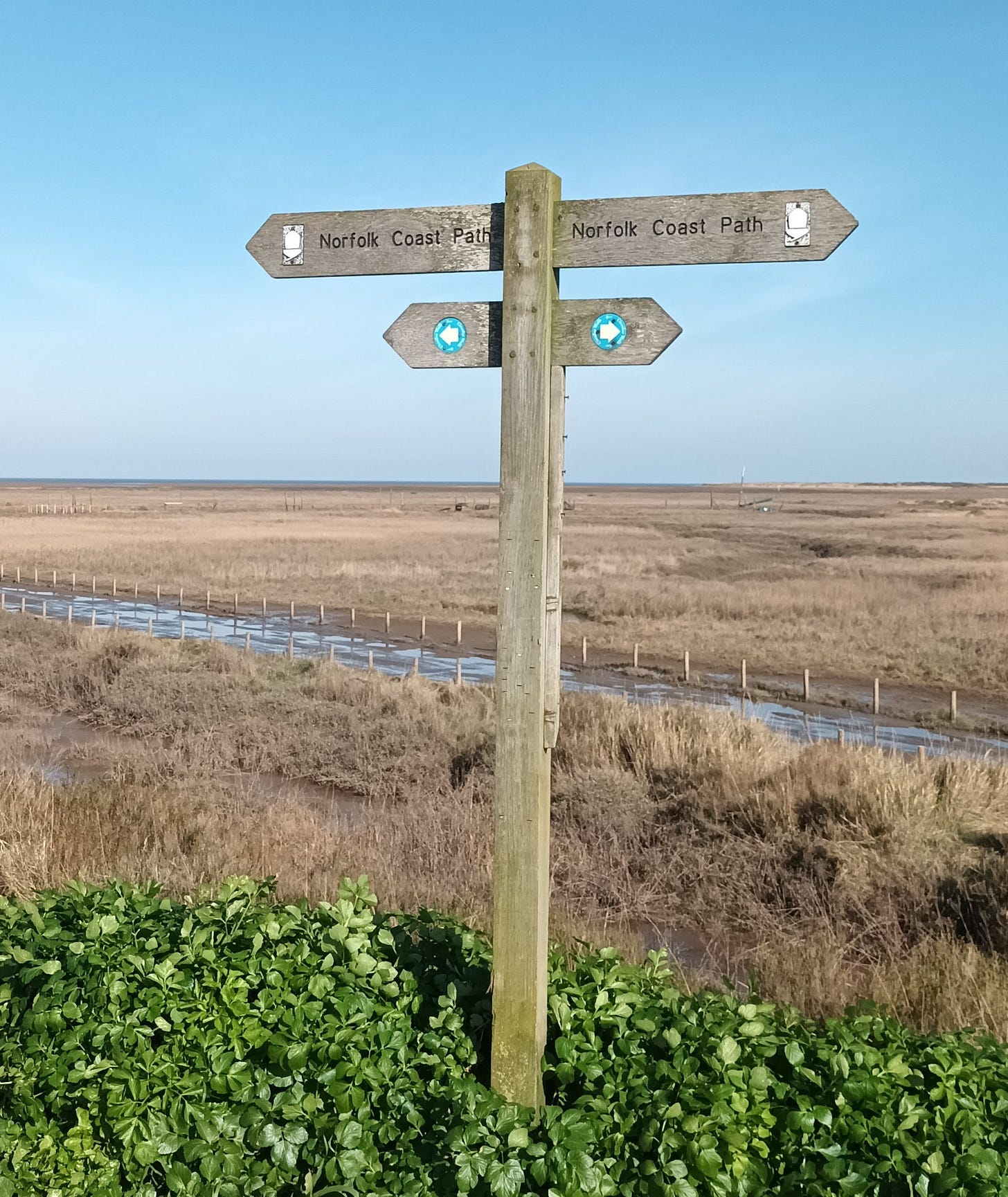 Footpath sign showing Norfolk Coast Path, marsh and blue sky behind