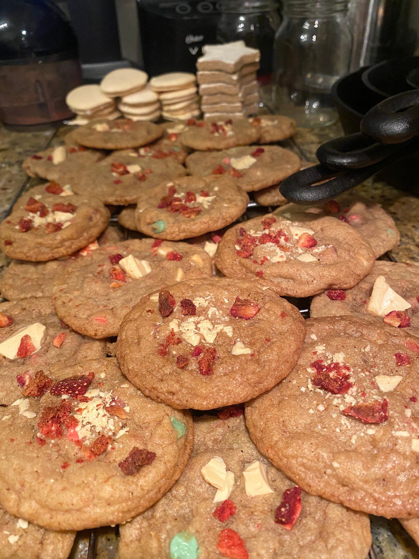 A cooling rack with stacks of the cookies described above, with white chocolate and dried strawberry pieces on top. In the background and stacks of star-shaped zimtsterne, and round meringues.