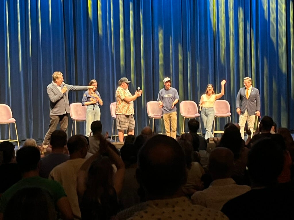 Six comedians stand in front of six chairs on a theater stage holding microphones and waving to the audiece. There are four men and two women. 