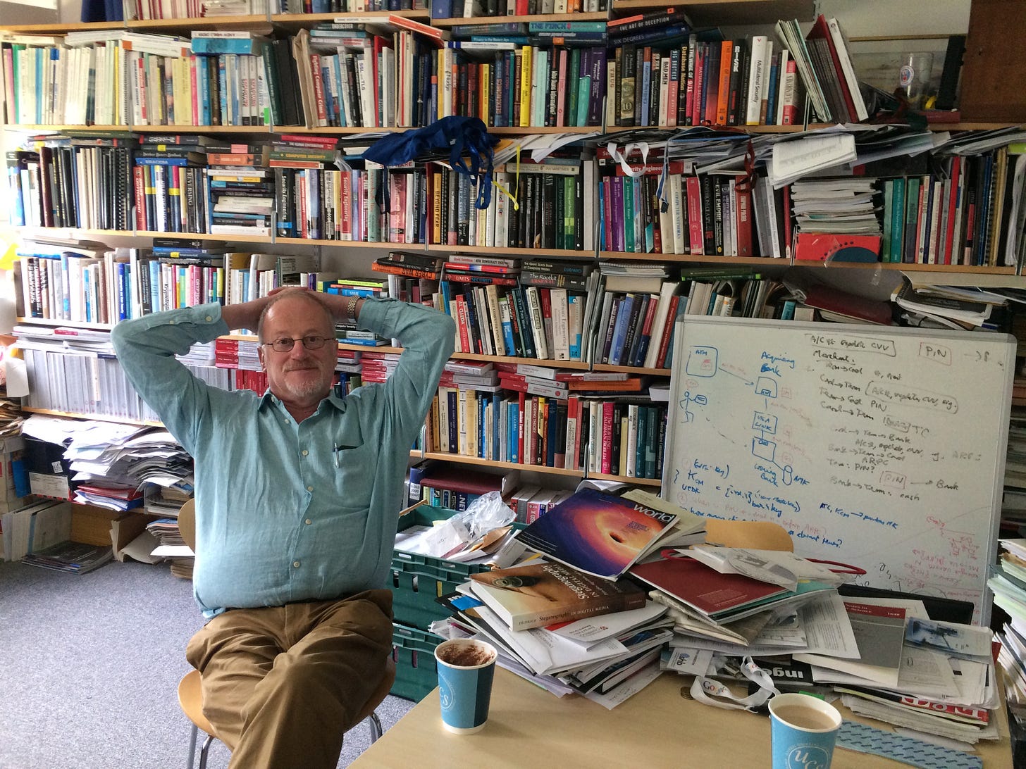 He is leaning back from a table with two cups of coffee/tea on it, with an enormous wall of books piled haphazardly on shelves behind him. There is also a whiteboard shown with equations.