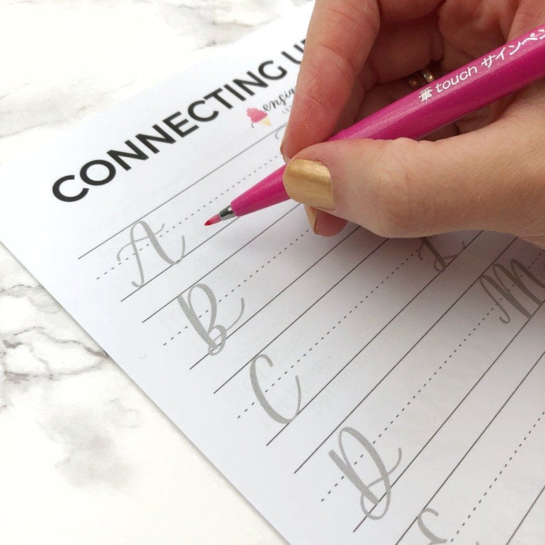 May include: A close-up of a hand using a pink brush pen to practice connecting cursive letters on a white sheet of paper. The paper has the words "CONNECTING UPPERCASE" printed at the top. The letters "A", "B", "C", and "D" are written in cursive on the paper.