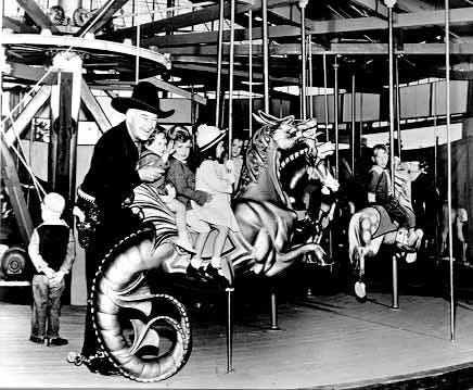 Photo of William Boyd in full Hopalong Cassidy regalia at Hoppyland in the early 1950s. He stands on a carousel supporting three small children who are all riding on the same sea horse.