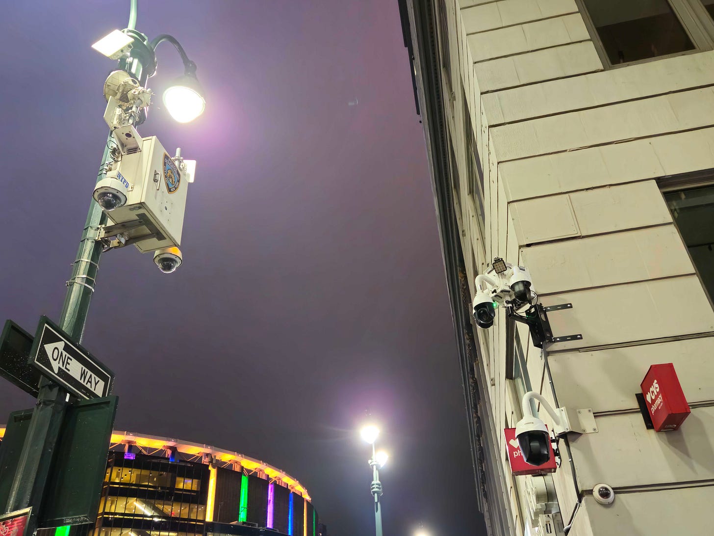 A photo of an NYPD surveillance camera on a light pole and a CVS surveillance camera on the corner of a building. A photo of an NYPD surveillance camera on a light pole and a CVS surveillance camera on the corner of a building.