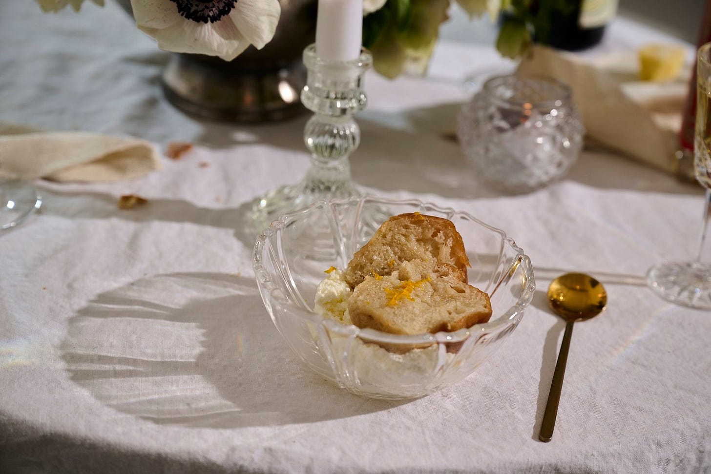 Baba au rhum served in a cut glass bowl with whipped cream and orange zest on a candlelit table set with flowers and vintage glassware.