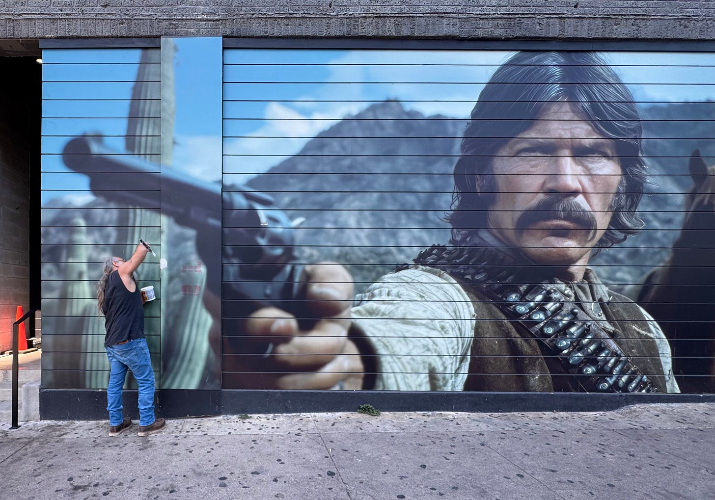 Man with ponytail, sleeveless black T-shirt and jeans patching bar exterior with huge mural of mustached cowboy resembling Charles Bronson aims a long revolver Man with ponytail, sleeveless black T-shirt and jeans patching bar exterior with huge mural of mustached cowboy resembling Charles Bronson aims a long revolver