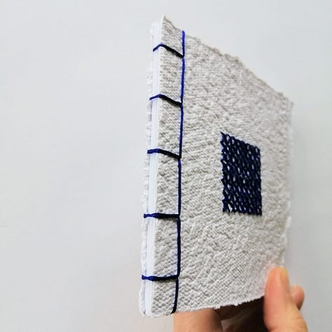 Handmade paper notebook bound using Japanese stab binding method with blue cross stitched square centre.