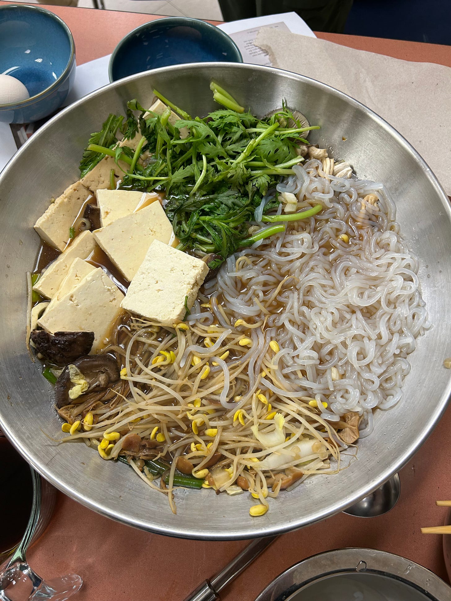 A wide metal pot containing tofu, chrysanthemum greens, shirataki, mushrooms, and bean sprouts in a bit of dark sauce. A wide metal pot containing tofu, chrysanthemum greens, shirataki, mushrooms, and bean sprouts in a bit of dark sauce.