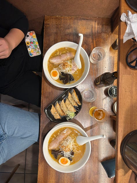 1. A aerial view of two bowls of miso ramen with char sui pork, half a soft boiled egg, wood ear mushrooms, and shredded green onions on top. In the middle of the bowls of ramen is a long plate of gyoza. 2. Brick building with entrance that says Sapporo Beer Musuem while snowing 3. Box on the street that has bags of sand that says in Japanese that people should feel free to scatter this sand to help everyone walk.