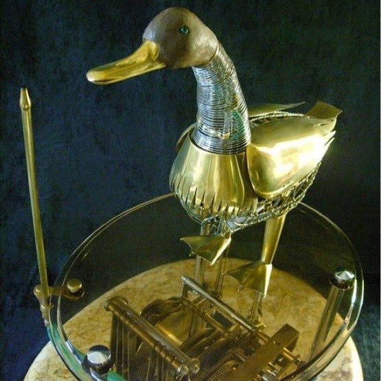 A largely brass duck, but with a wooden-looking head and springy neck, standing on a glass disc, with a mechanism underneath