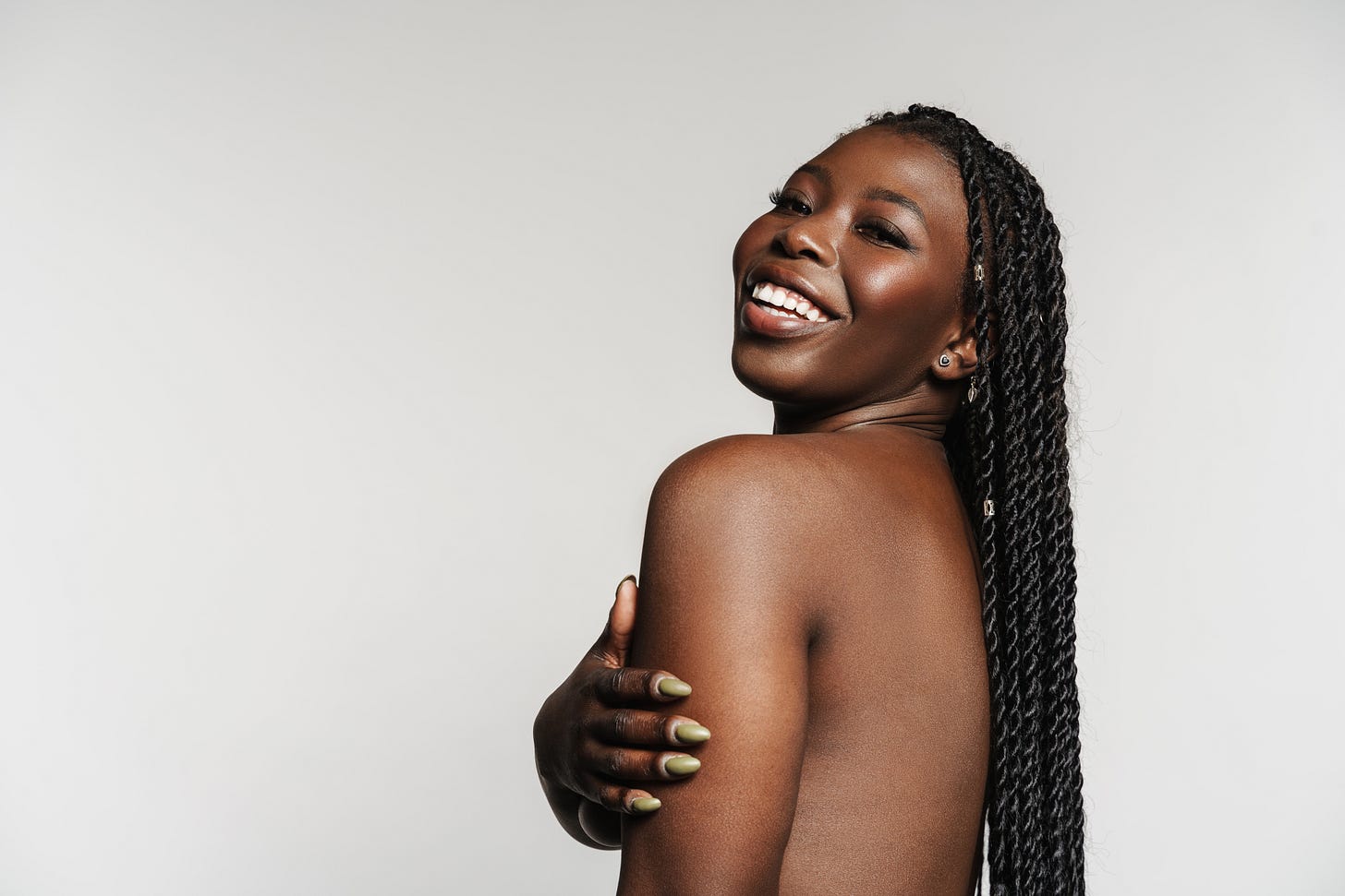 A smiling Black woman with long braided hair, nude from the shoulders up, looking over her shoulder with confidence and joy against a plain studio background. A smiling Black woman with long braided hair, nude from the shoulders up, looking over her shoulder with confidence and joy against a plain studio background.