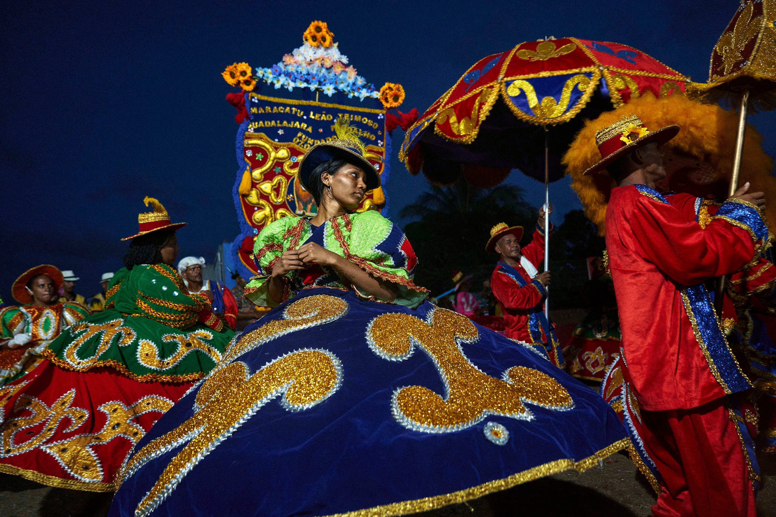 A woman in a vibrant, wide, blue and gold costume dances during a colorful, nighttime festival parade, surrounded by other performers in elaborate, brightly decorated outfits and large hats.