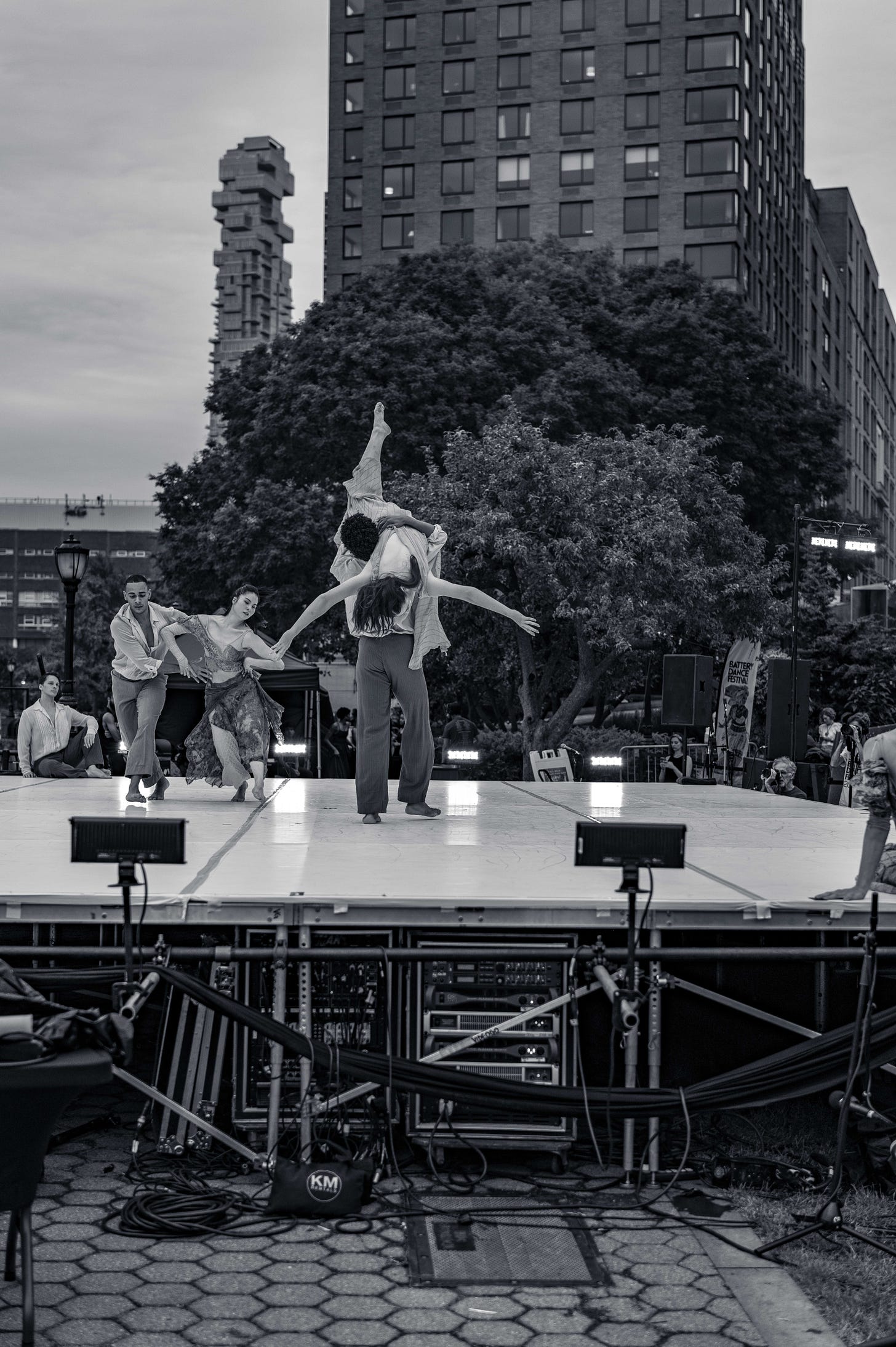 A group of contemporary dancers performing on an outdoor stage in Battery Park, with one dancer lifted high against a backdrop of trees and city buildings. A group of contemporary dancers performing on an outdoor stage in Battery Park, with one dancer lifted high against a backdrop of trees and city buildings.