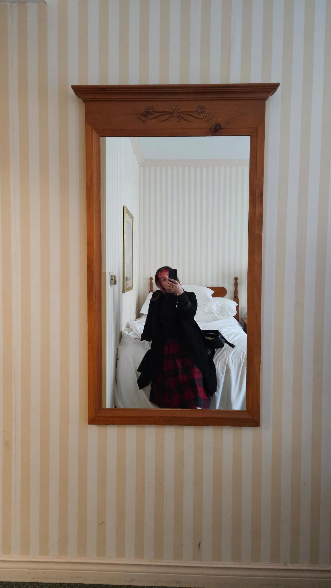 A mirror with a light brown frame is mounted on a wall with stripes of pale yellow and white. A room with a white bed is reflected and Murph sits on the bed in a black wool coat and tartan skirt, taking a selfie