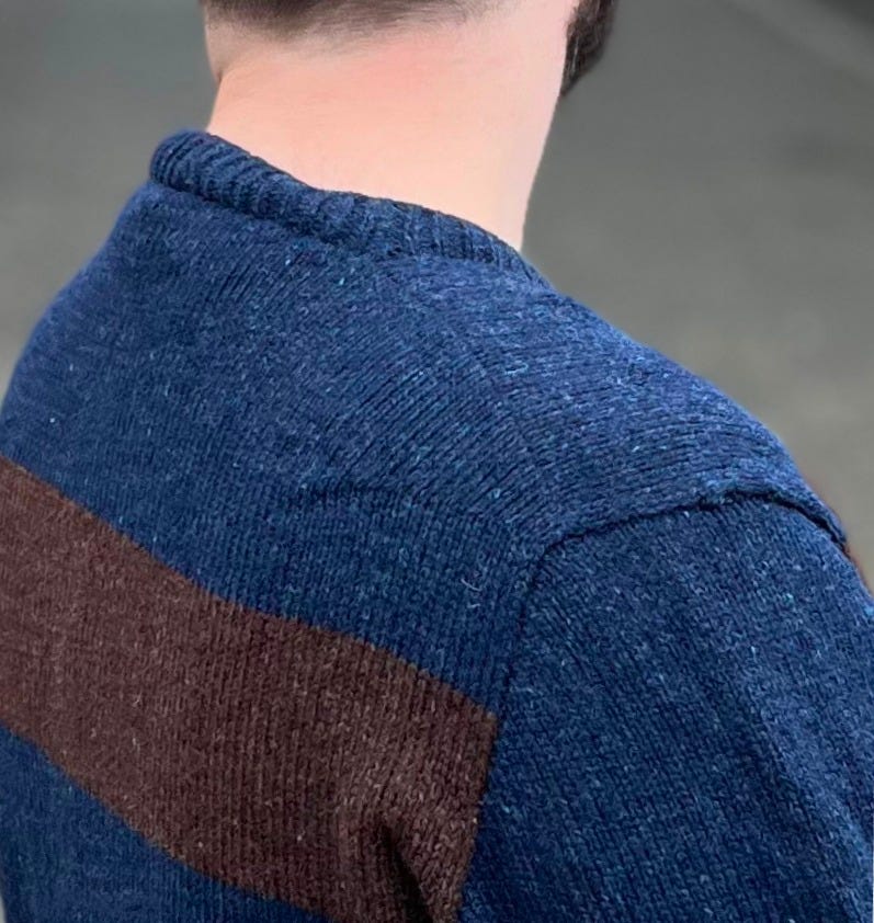 Close-up of the back of a knitted sweater at the shoulder, showing the ribbed collar and the shoulder seam set behind the top of the shoulder, navy above and chocolate brown stripe below.