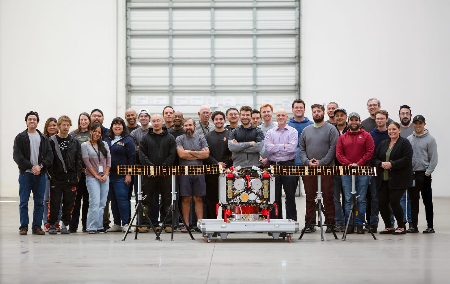 A group of people poses indoors behind a robotic device with extended solar panels, positioned in front of a large, closed industrial door. A group of people poses indoors behind a robotic device with extended solar panels, positioned in front of a large, closed industrial door.
