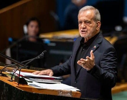 Masoud Pezeshkian gestures with one hand while speaking from a lectern with microphones and papers. A blurry assembly hall is in the background.