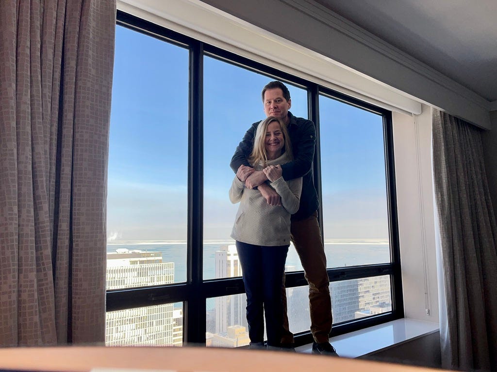 A tall, brown-haired man hugs a short blonde woman. They're standing in front of a tall window overlooking Lake Michigan.