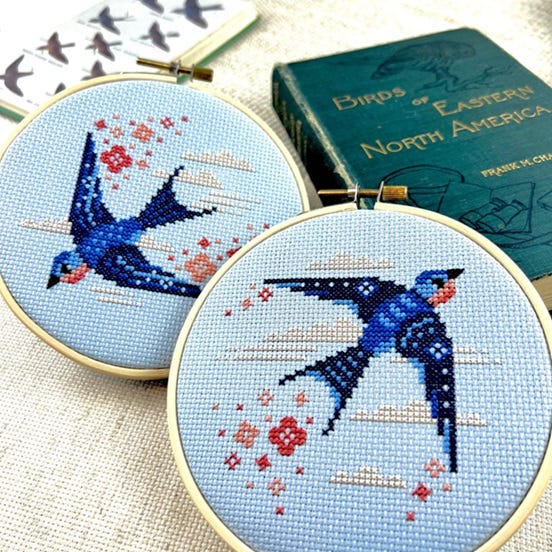 Two hooped cross stitched project are staged with a vintage edition of 'Birds of Eastern North America.' The cross stitched projects are of Swallows, in deep blue hues with subtle floral and cloud motifs.