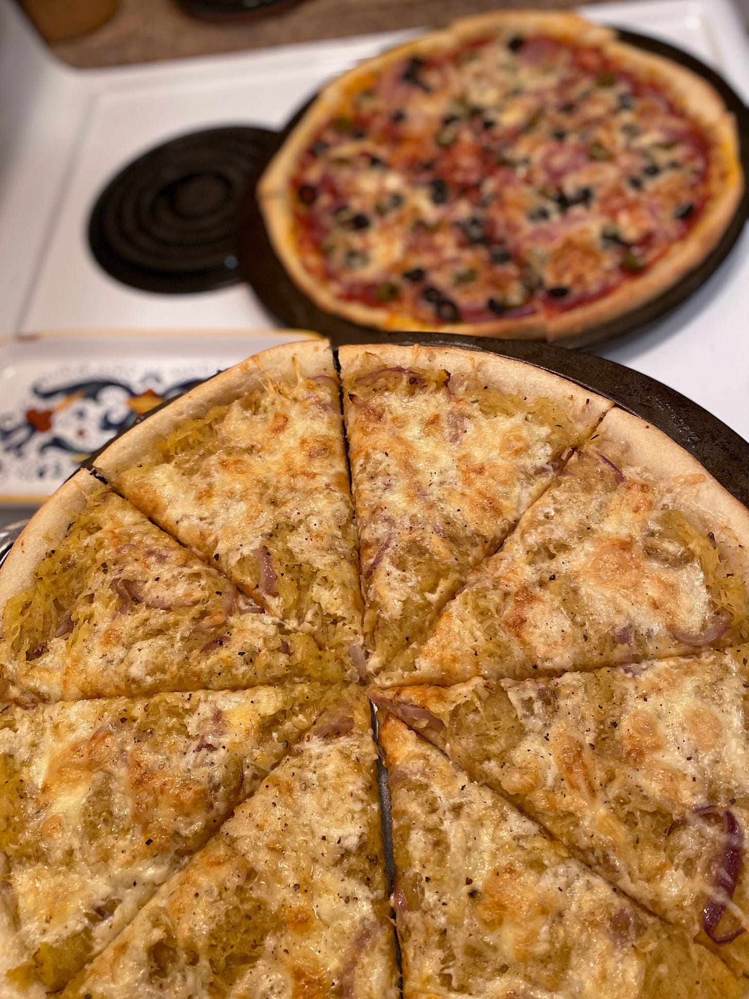 Two pizzas in pans on top of the stove. In the foreground is the spaghetti squash one, and the tomato base one with olives is behind it.
