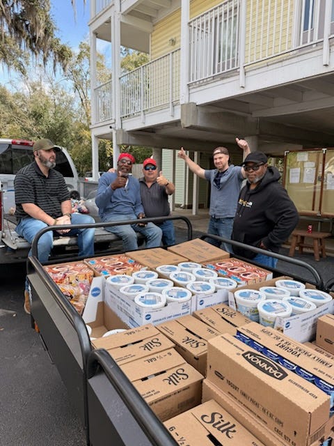 Five men pose outdoors beside a pickup truck and a trailer loaded with boxes and cartons of food, including multiple “Silk” and “Dannon” boxes, in front of a raised yellow building with white railings and a staircase. Some of the men give thumbs-up and raise their arms, and there are tables and stacked pallets in the background under the building.