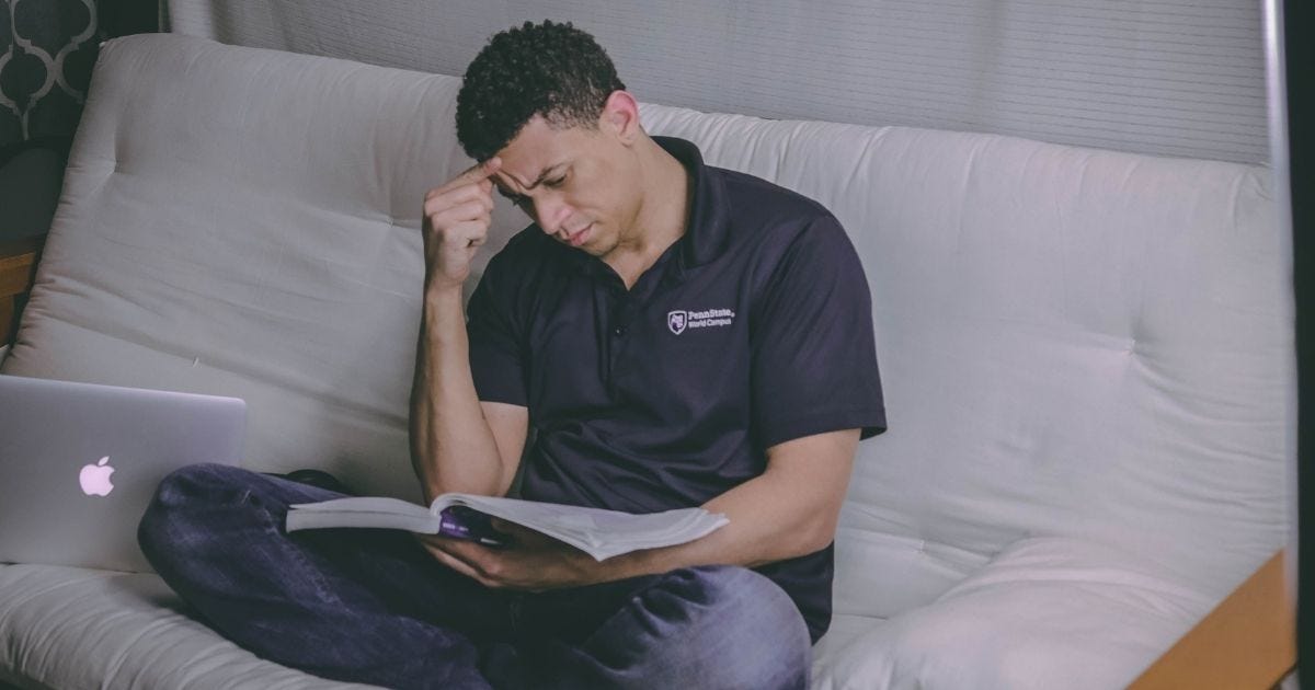 A student relaxing on a sofa, studying with a book and using a laptop indoors A student relaxing on a sofa, studying with a book and using a laptop indoors