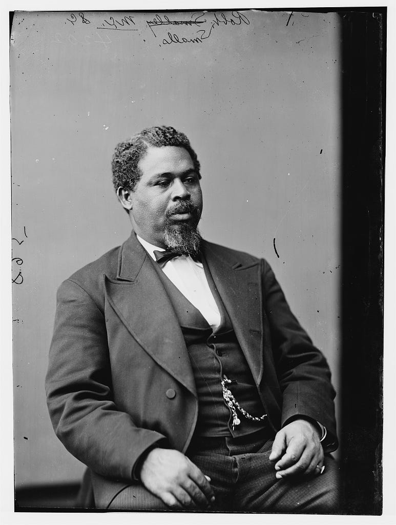 Black-and-white studio portrait of Robert Smalls seated in formal nineteenth-century dress, wearing a dark suit, vest, bow tie, and watch chain, with archival handwriting visible along the top edge. Black-and-white studio portrait of Robert Smalls seated in formal nineteenth-century dress, wearing a dark suit, vest, bow tie, and watch chain, with archival handwriting visible along the top edge.