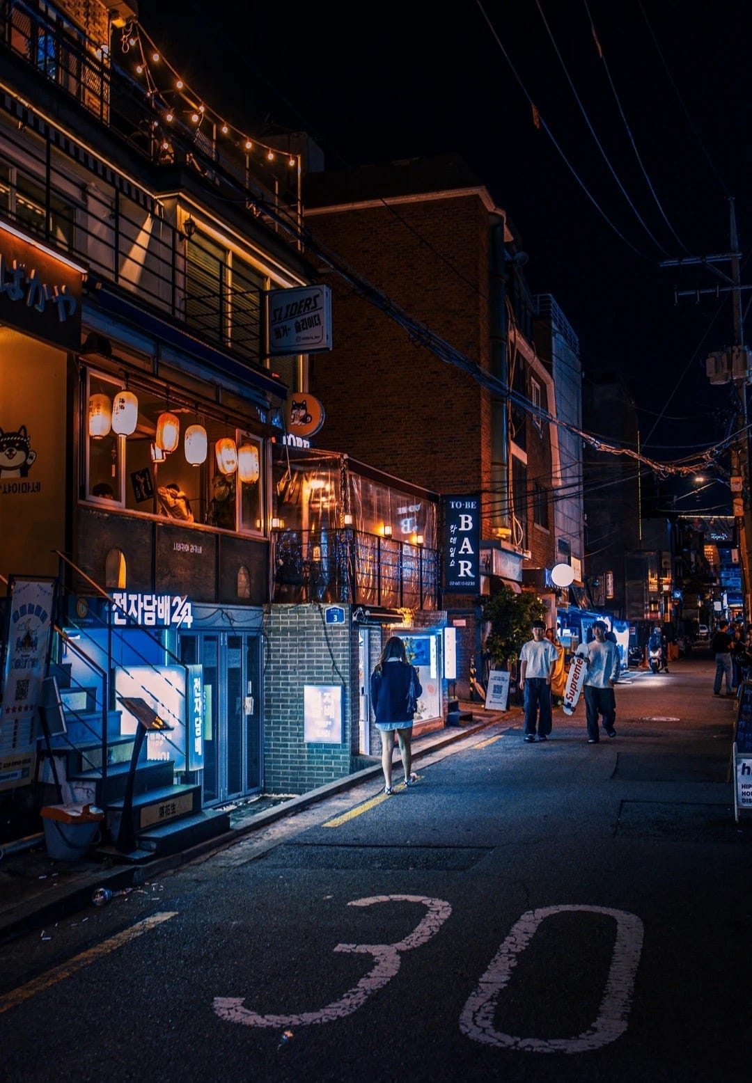 A narrow Seoul side street at night, lit by warm paper lanterns and neon Korean bar signs, with a few people walking past small izakaya-style bars and a painted "30" speed marking on the road.
