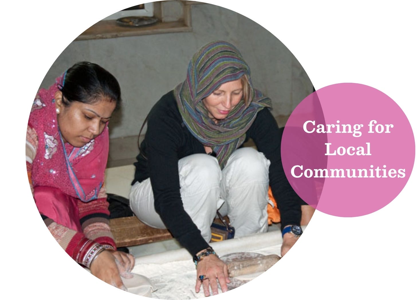 A South Asian woman wearing a pink headscarf and a white woman wearing a striped head scarf are kneeling together rolling our bread for naan.