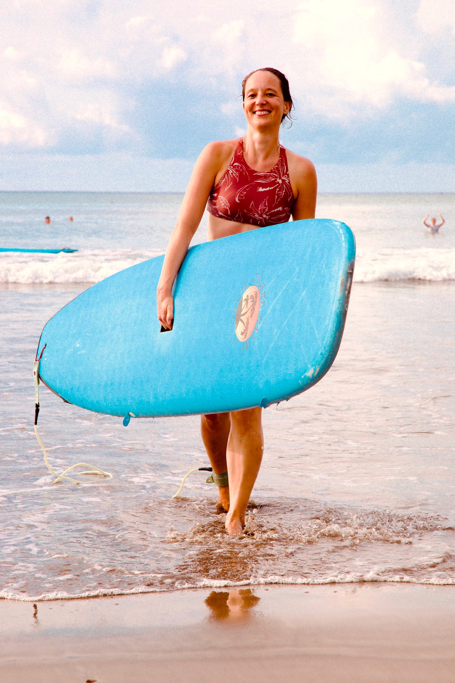 Woman with ponytail in red patterned swimsuit walking on beach while carrying blue surfboard, smiling broadly with ocean and cloudy sky in background. Photograph by Georges Yazbek.