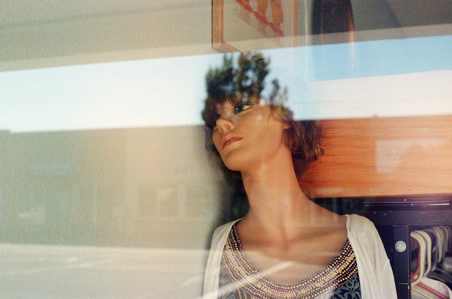A film photograph of a female mannequin leaning against a wall, as if despondently. The camera is looking through a glass window, so color & light reflections can be seen accordingly.