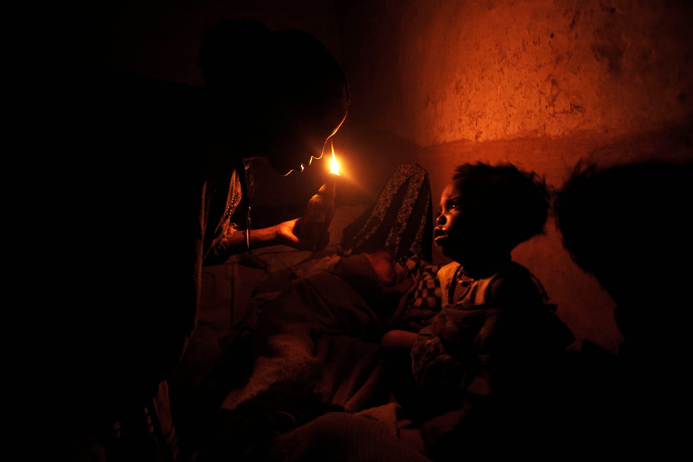 A person holds a small candle near a young child in a dimly lit, rough-walled room, casting a warm glow on the child’s face as they look up at the flame.