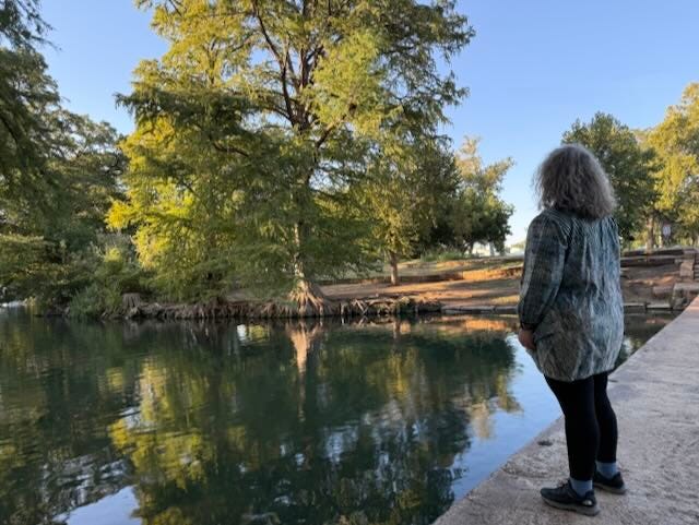 Woman on riverbank looking up at tall cypress trees with water reflections