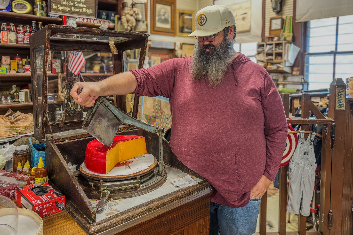 Ein bärtiger Mann mit Baseballkappe steht in einem historischen Laden in Harrison County, Texas, und bedient eine alte manuelle Käseschneidemaschine, mit der er ein grosses Stück rot-gelb gewachsten Käse schneidet; um ihn herum Regale mit Vintage-Produkten, Schildern und Alltagsgegenständen.