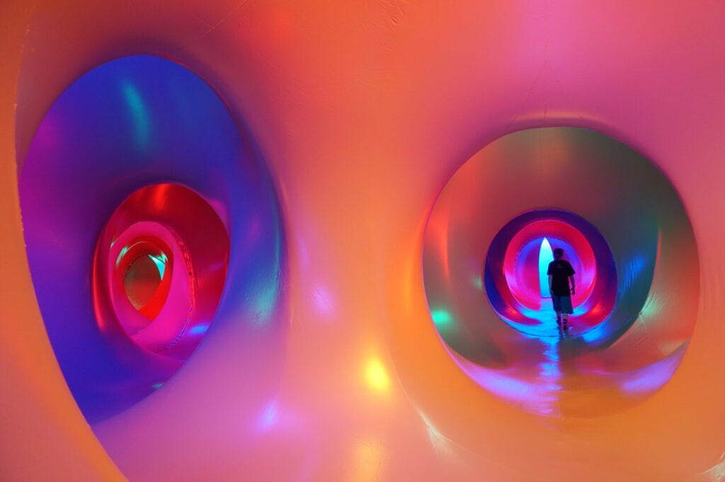 We are looking at the interior of a Luminarium, an inflatable structure with corridors and chambers bathed in coloured light. From this view we are standing in the intersection of two corridors that can both be seen down. The corridors are circular and oval in shape and undulate side to side as they progress. The foreground where we are is a soft peach colour, and you can see the colours morph and blend as your eye travels down the corridors into blues, pinks and reds with purple and green spotlights at points adding pattern. You can see the black silhouette of a lone figure walking down the end of corridor on the right.