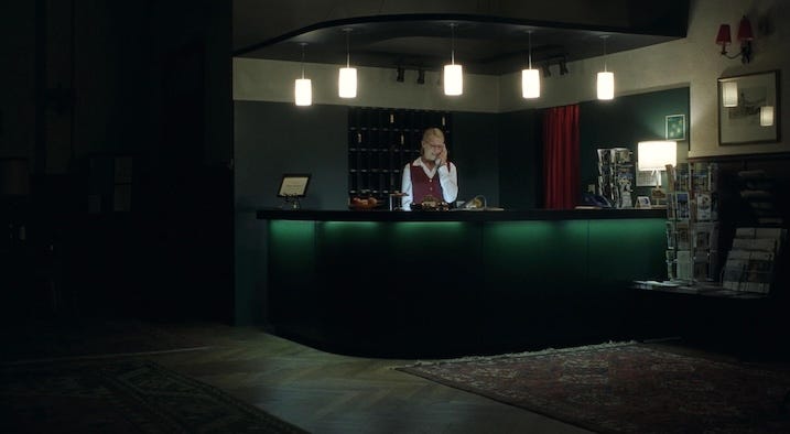 A hotel receptionist in uniform stands behind a dimly lit front desk, speaking on the phone. The setting is formal but unsettling, with deep shadows and harsh green lighting.