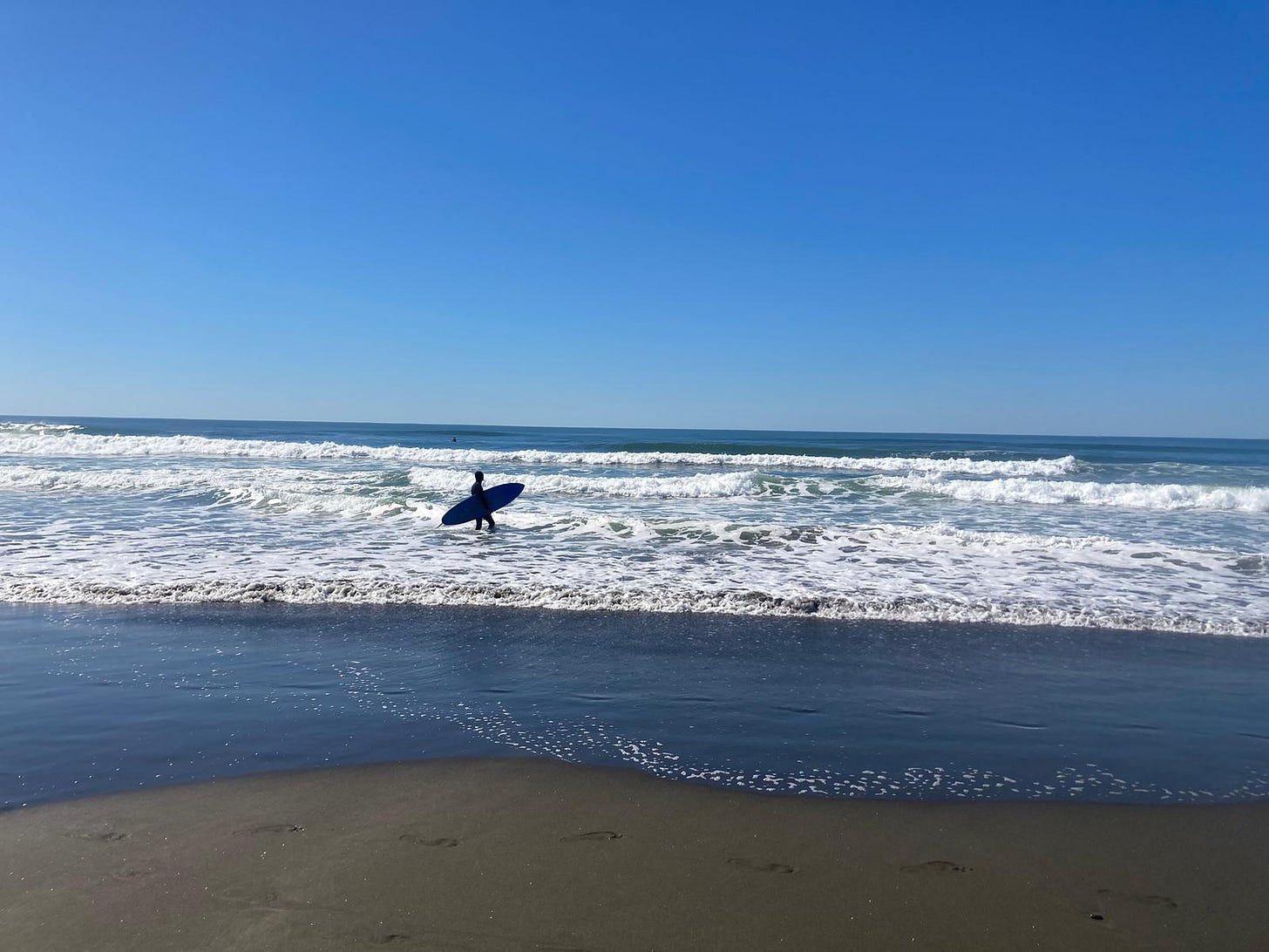 surfer stands in the white surfbreak with sand in the foreground.