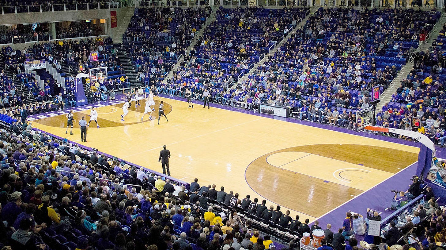 Crowded basketball game in a large indoor arena. Players in action on court. Purple and blue seats filled with spectators. Excited atmosphere.