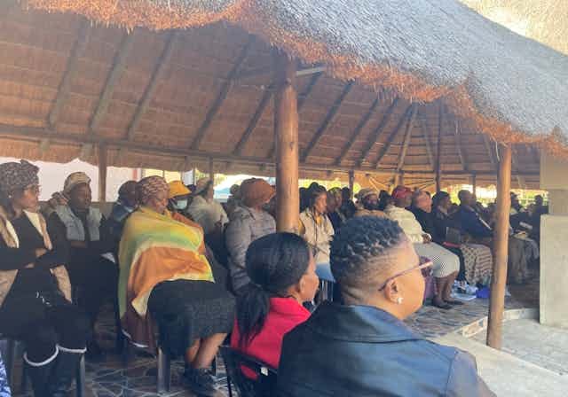 A group of people sit under a thatched roof. A group of people sit under a thatched roof.