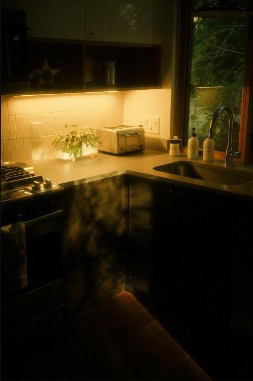 An empty kitchen at dusk, warm light glowing under the cabinets, a dark window looking out onto trees. An empty kitchen at dusk, warm light glowing under the cabinets, a dark window looking out onto trees.
