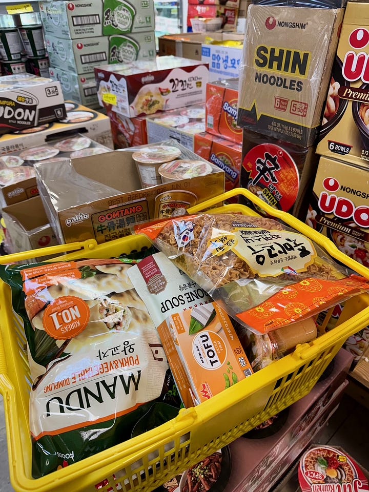 can of lee kum king chicken bouillon powder, full hand cart at korean grocery store, pot of korean noodles and green vegetables, mushrooms and chicken, hand-painted ramen sign hangs above shelves of ramen
