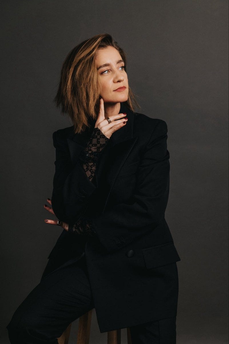 A woman with shoulder-length wavy brown hair and a thoughtful expression sits on a wooden stool against a dark gray background. She wears a black blazer over a sheer black lace top, with her right hand raised to her chin in a pondering pose, fingers touching her lips. Her left arm rests on her lap, and she has a silver ring on her right index finger. The outfit includes black pants, and the overall style is professional.