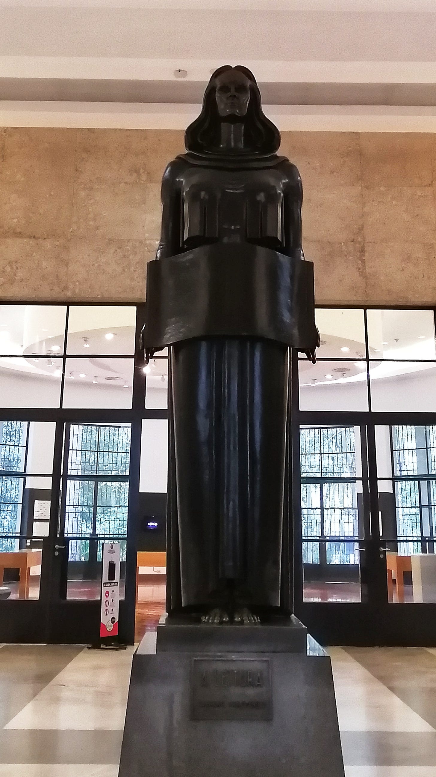Estátua em metal escuro de mulher em pé segurando um livro aberto a frente do corpo.
