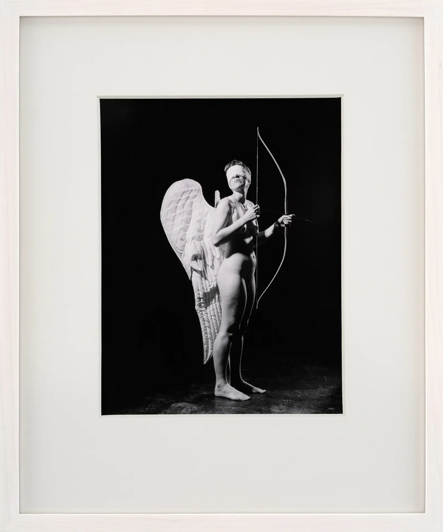 Black and white photo of a nude white woman with large white angel wings holding a bow, resembling a mythical figure. Framed against a dark background, evoking a serene, ethereal mood. The photo is in a white matte, with a white frame around it, hung on a white wall. The person is wearing a white blindfold over her eyes and she is looking up and to her right.