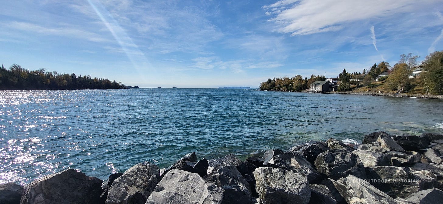 A view from a pier looking down the shoreline. Small cottages line the water's edge to the right, protected by rocky breakwalls. The vast, open lake extends to the left.