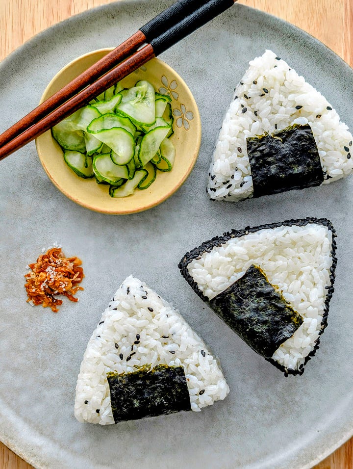 Plate of three mushroom pâté-filled onigiri with pickled cucumbers and crispy onion salt next to a plate of two pickled cherry blossom onigiri, showcasing different Japanese rice ball styles.