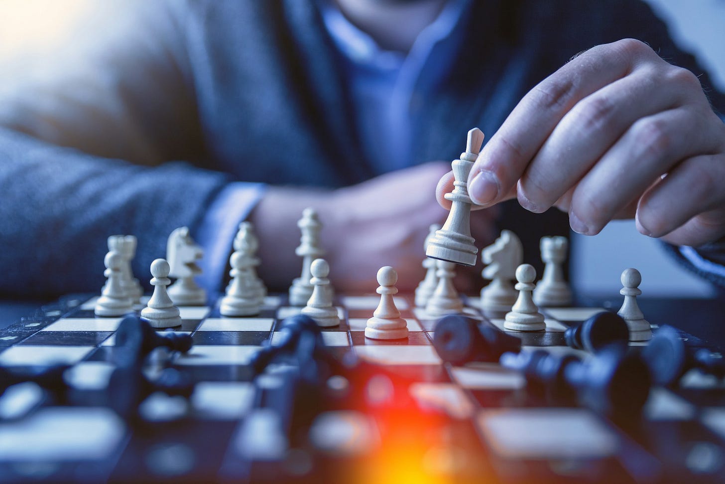 A man sits at a chessboard, his face out of shot. He holds up a white piece, moving it over black pieces that have been knocked down to a final move. A man sits at a chessboard, his face out of shot. He holds up a white piece, moving it over black pieces that have been knocked down to a final move.