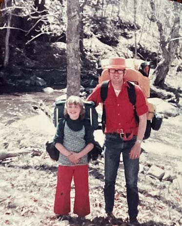 girl and dad with backpacks on in front of stream, two women by trailhead sign in forest, solo woman beside mossy trail