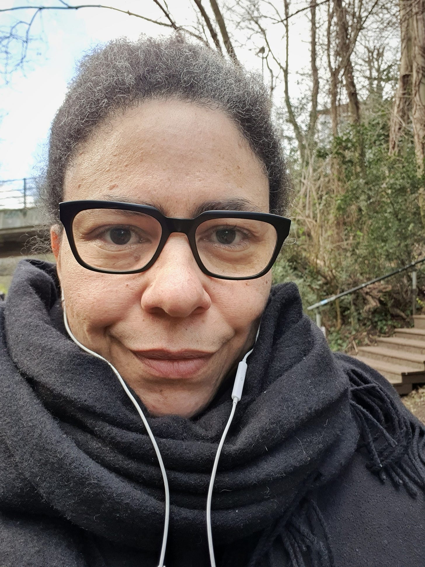 Close up photo of a woman outdoors, she is wearing glasses and has a scarf wrapped round her neck