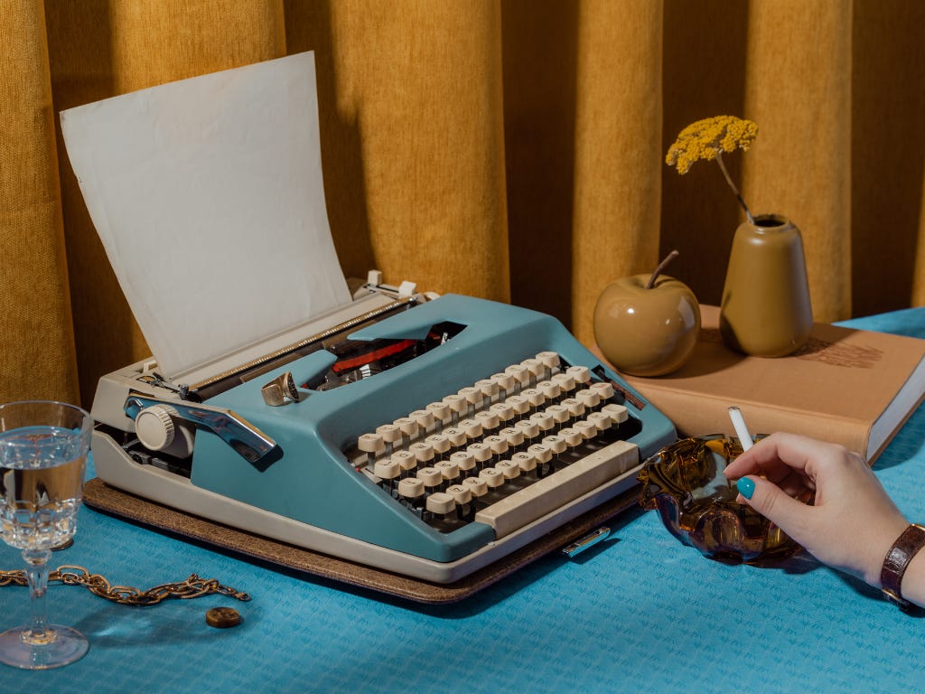 Blauwe retro typemachine op een turquoise tafel met een hand naast een asbak, boek, glas en gele bloemen. Blauwe retro typemachine op een turquoise tafel met een hand naast een asbak, boek, glas en gele bloemen.